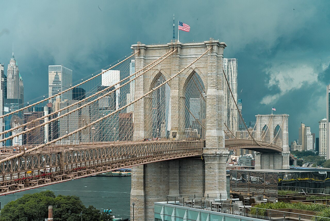 A Brooklyn Bridge tower under storm clouds, the Chrysler and Midtown behind