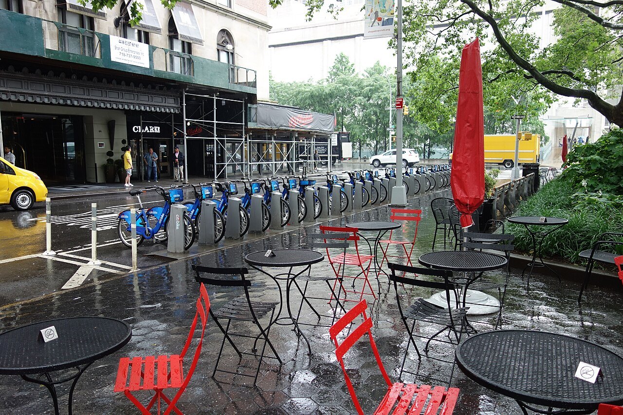 Citi Bikes docked beside café tables on a wet day at Dante Park
