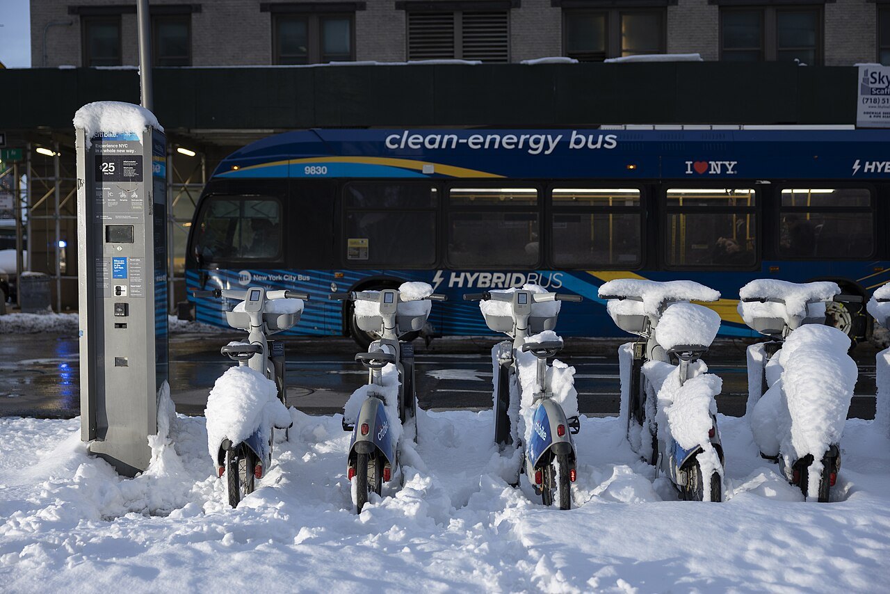 Snow-covered Citi Bikes at a dock as a bus passes