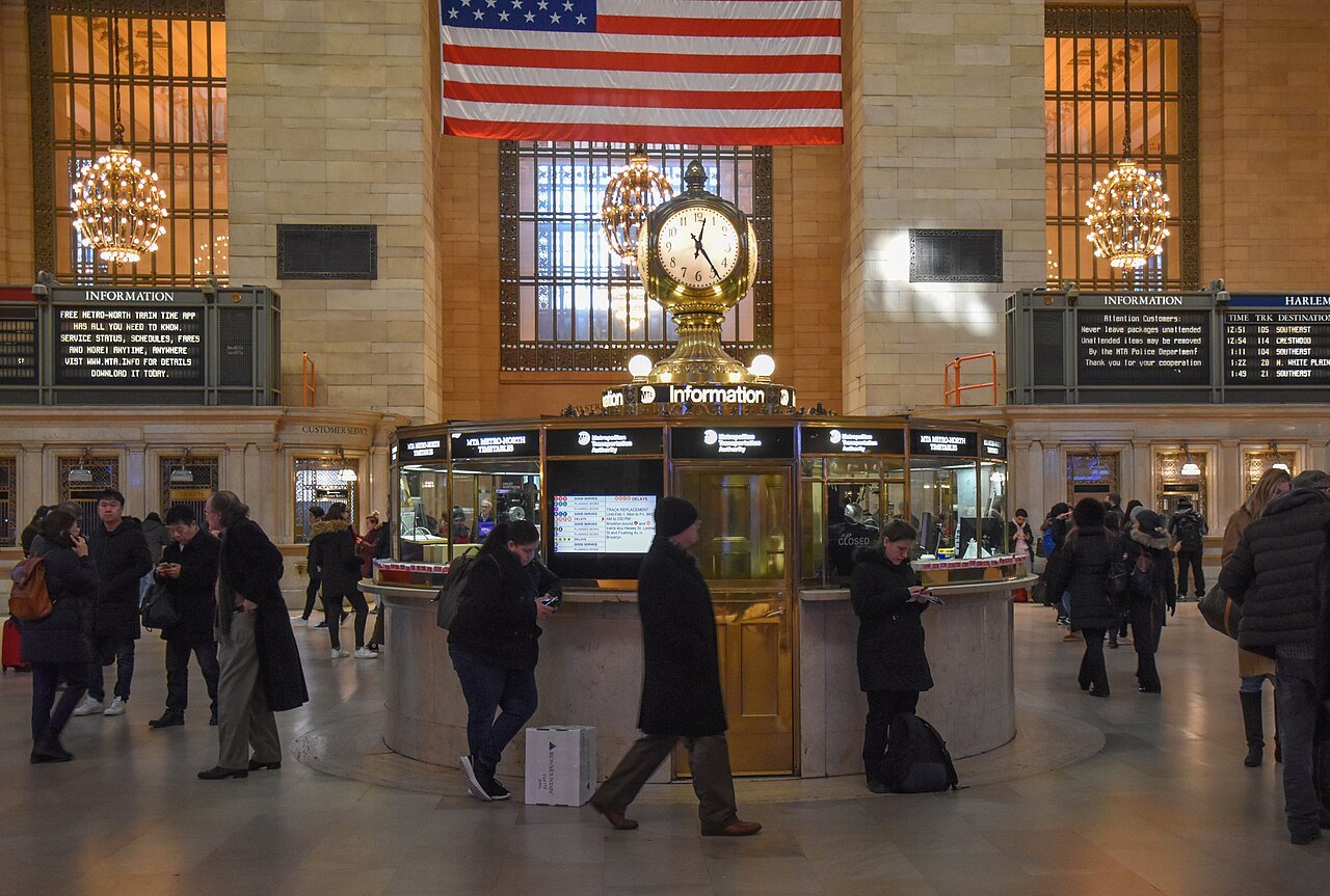 The brass four-faced clock above the information booth, the flag beyond