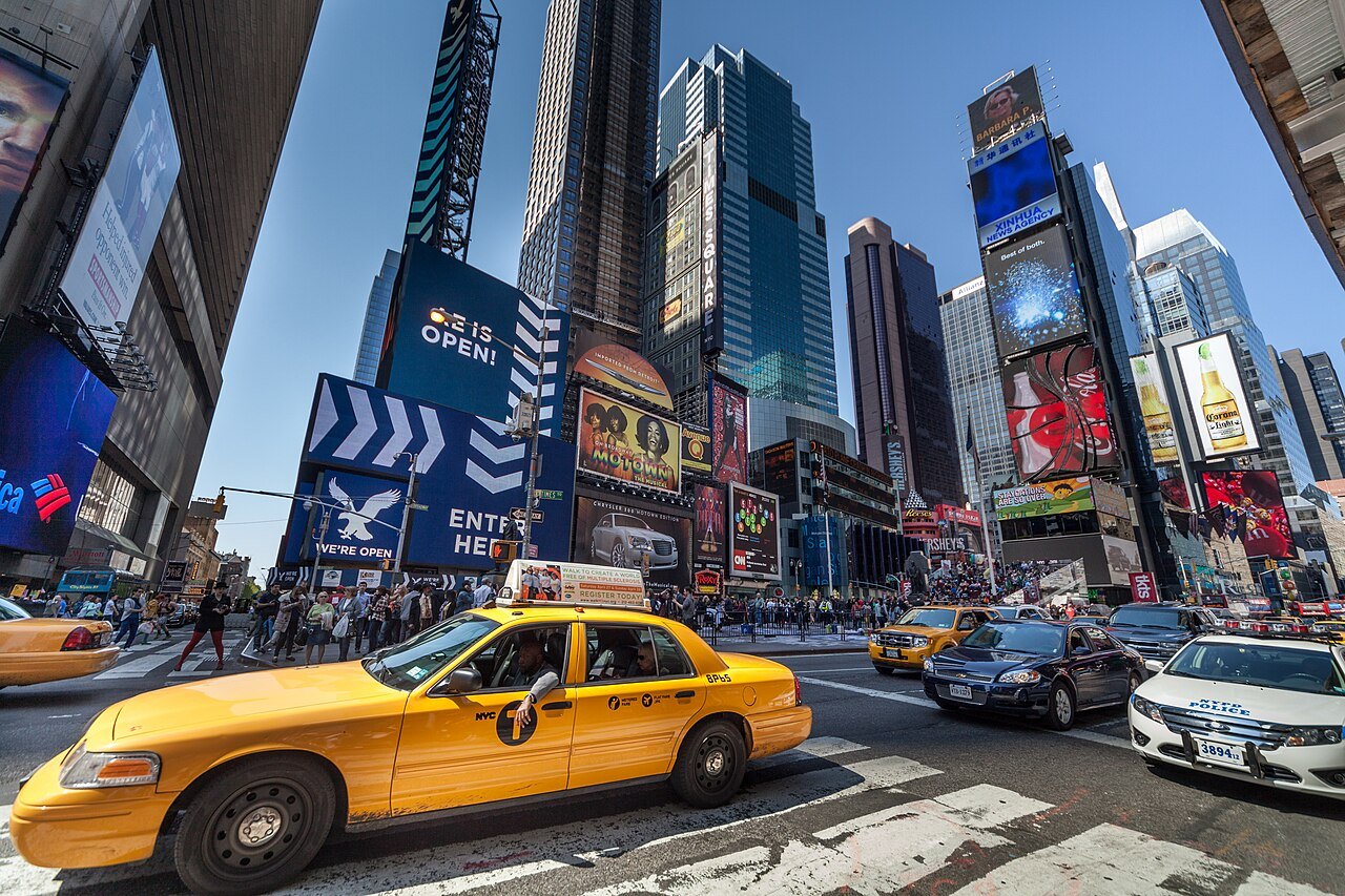 A yellow cab passing the towers and screens of the Theater District